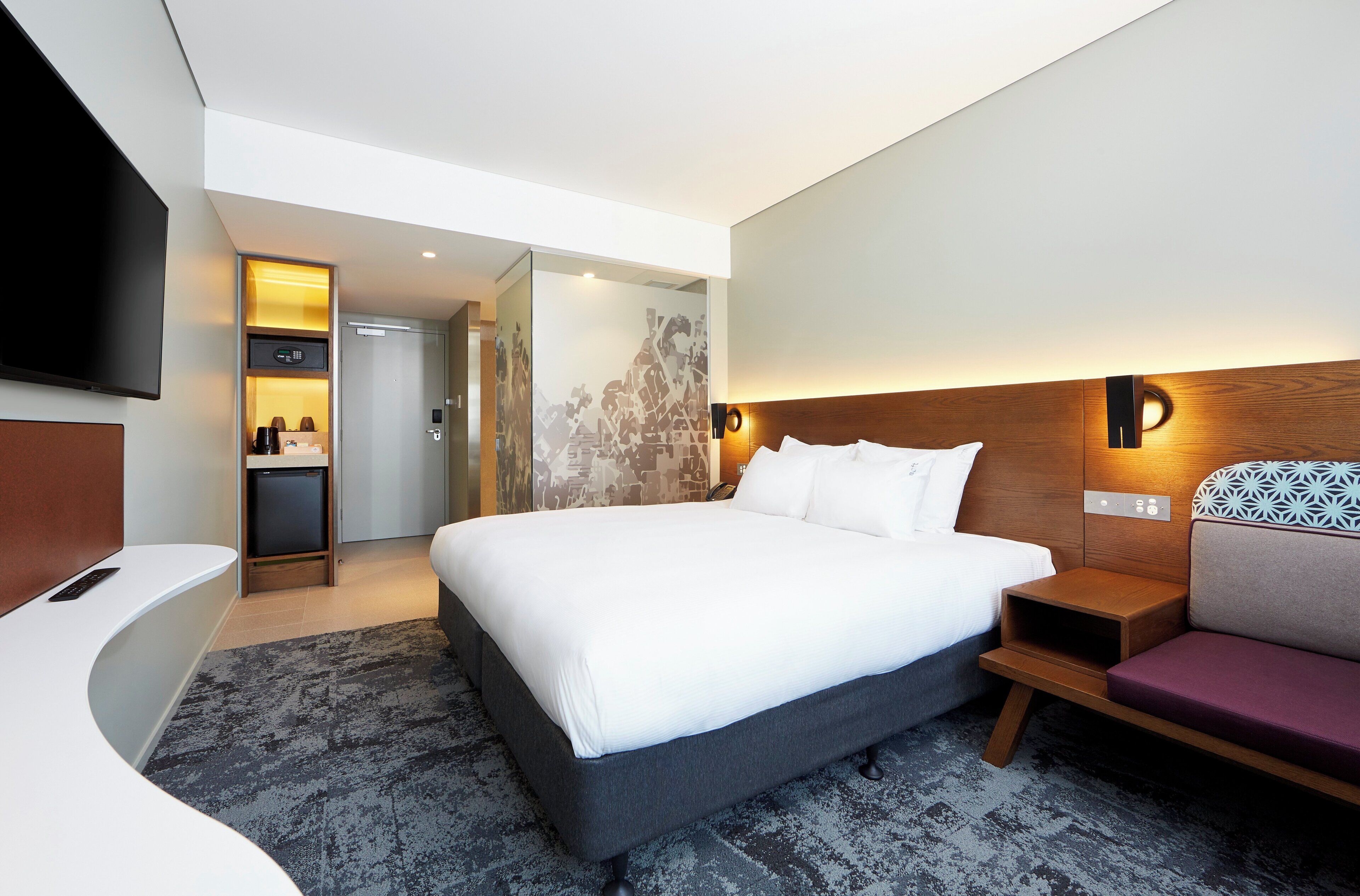 A modern hotel room with a large bed, a seating area, a TV, and a small kitchenette.