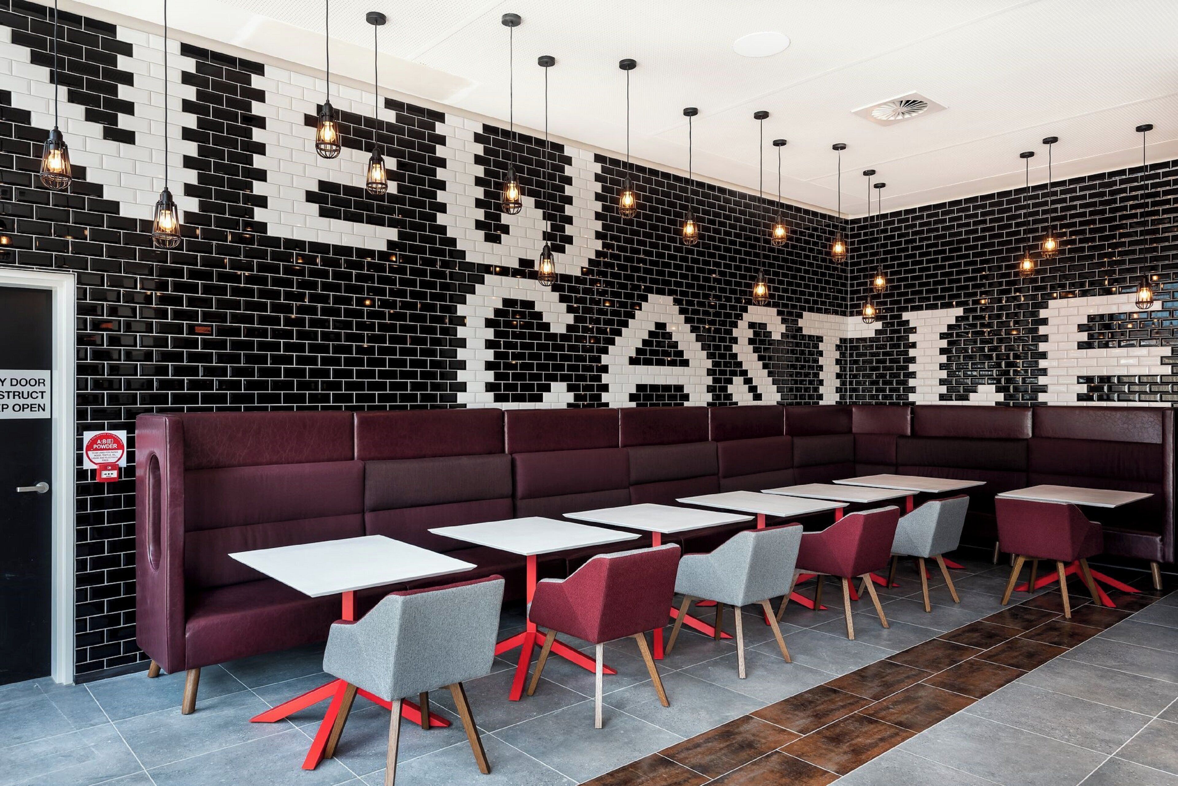 A restaurant interior with a black and white tiled wall, red cushioned booths, and grey and red chairs.