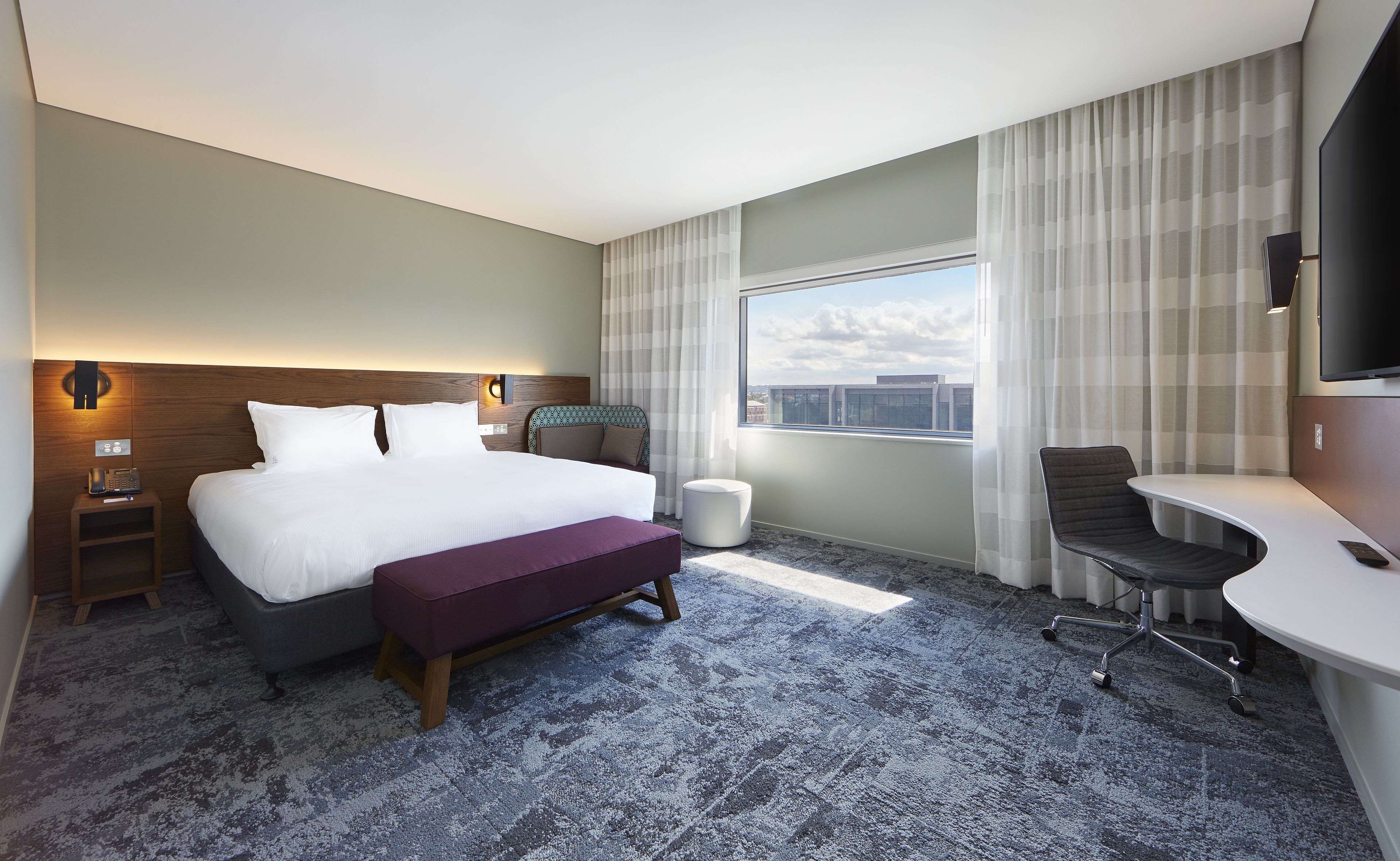 Een hotelkamer met een groot bed, een bureau, een stoel, een klein voetenbankje en uitzicht op de stad door het raam.