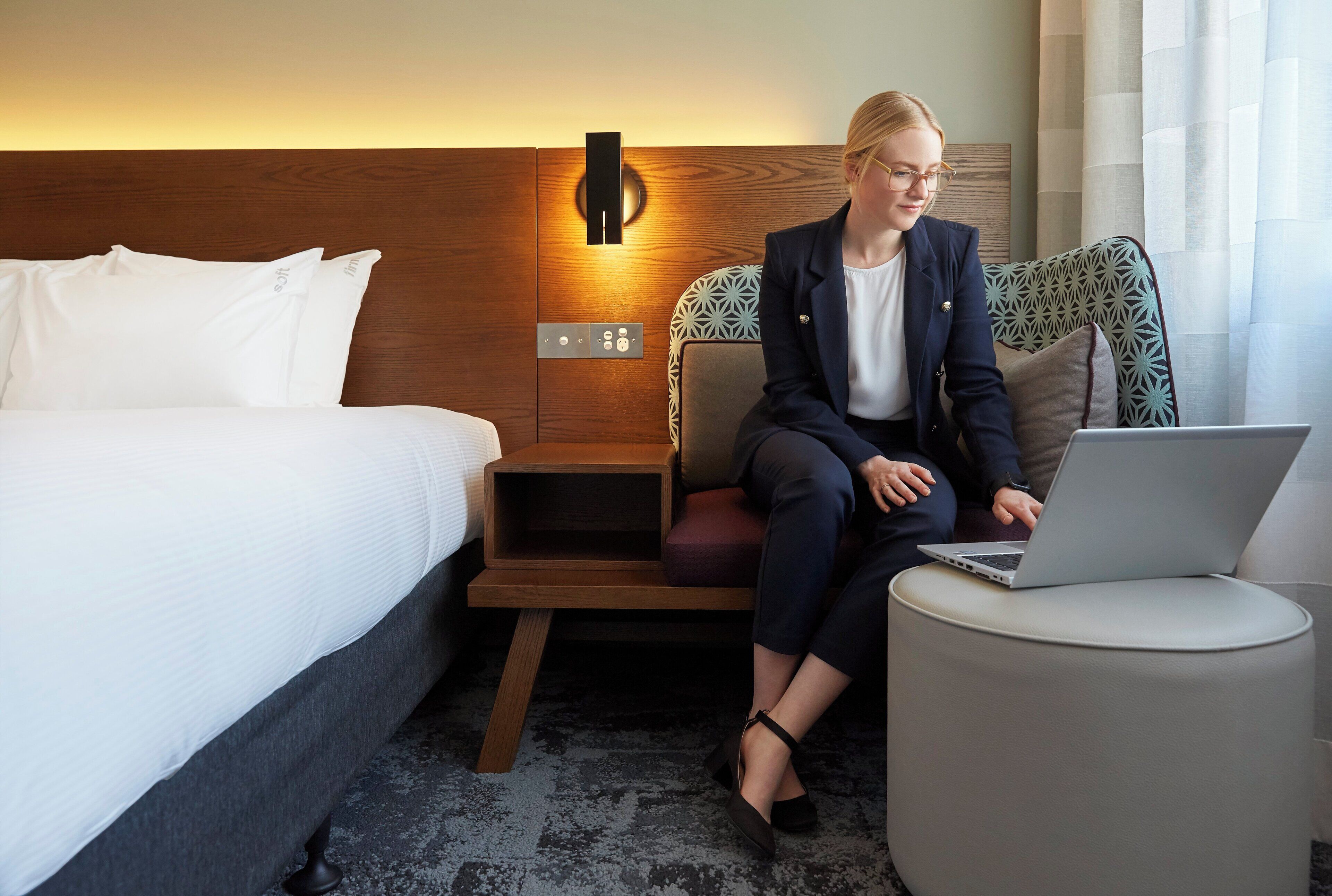 A hotel room with a bed, a desk, and a chair. There is a laptop on the desk.