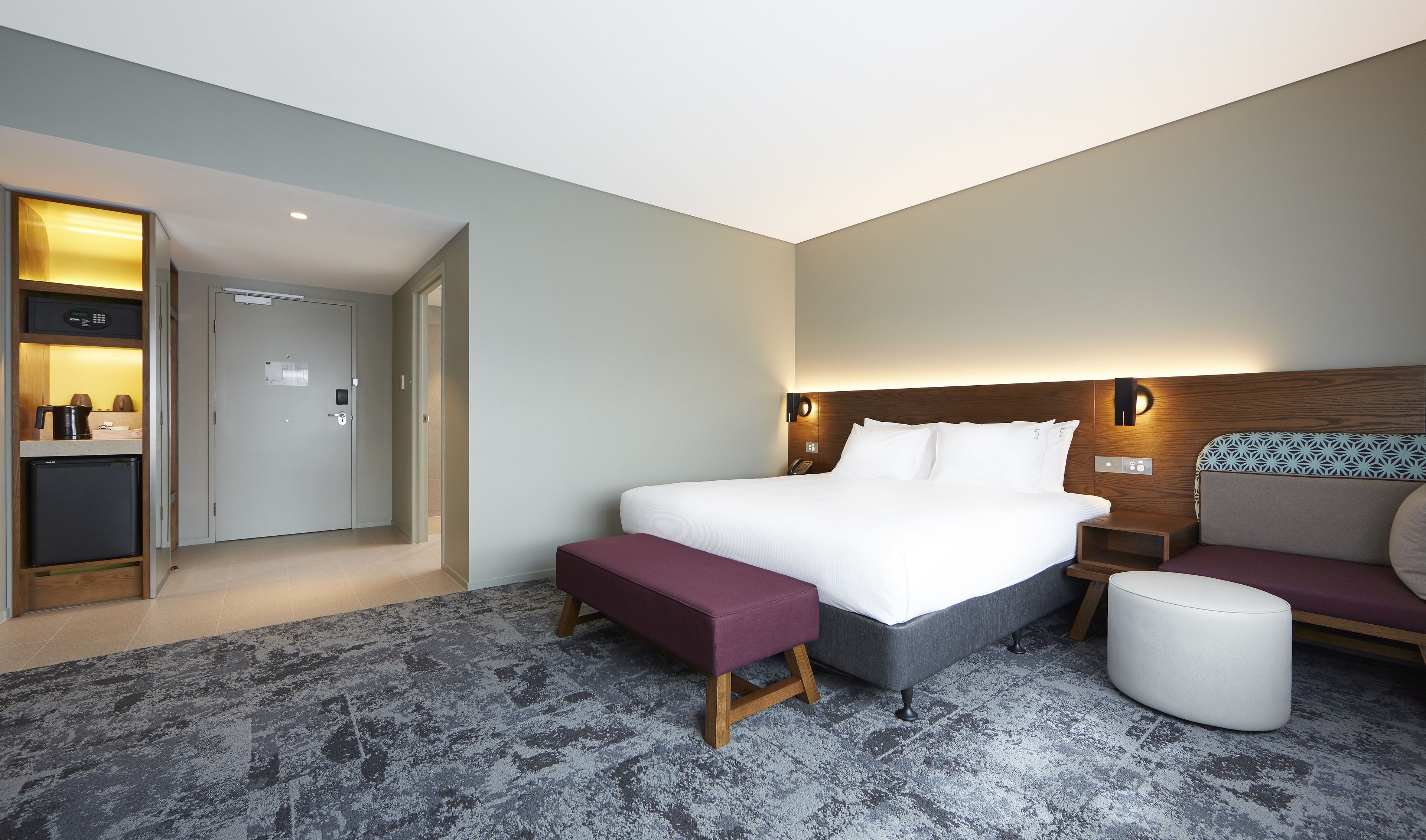 A modern hotel room with a large bed, a bedside table, a small seating area, and a kitchenette.