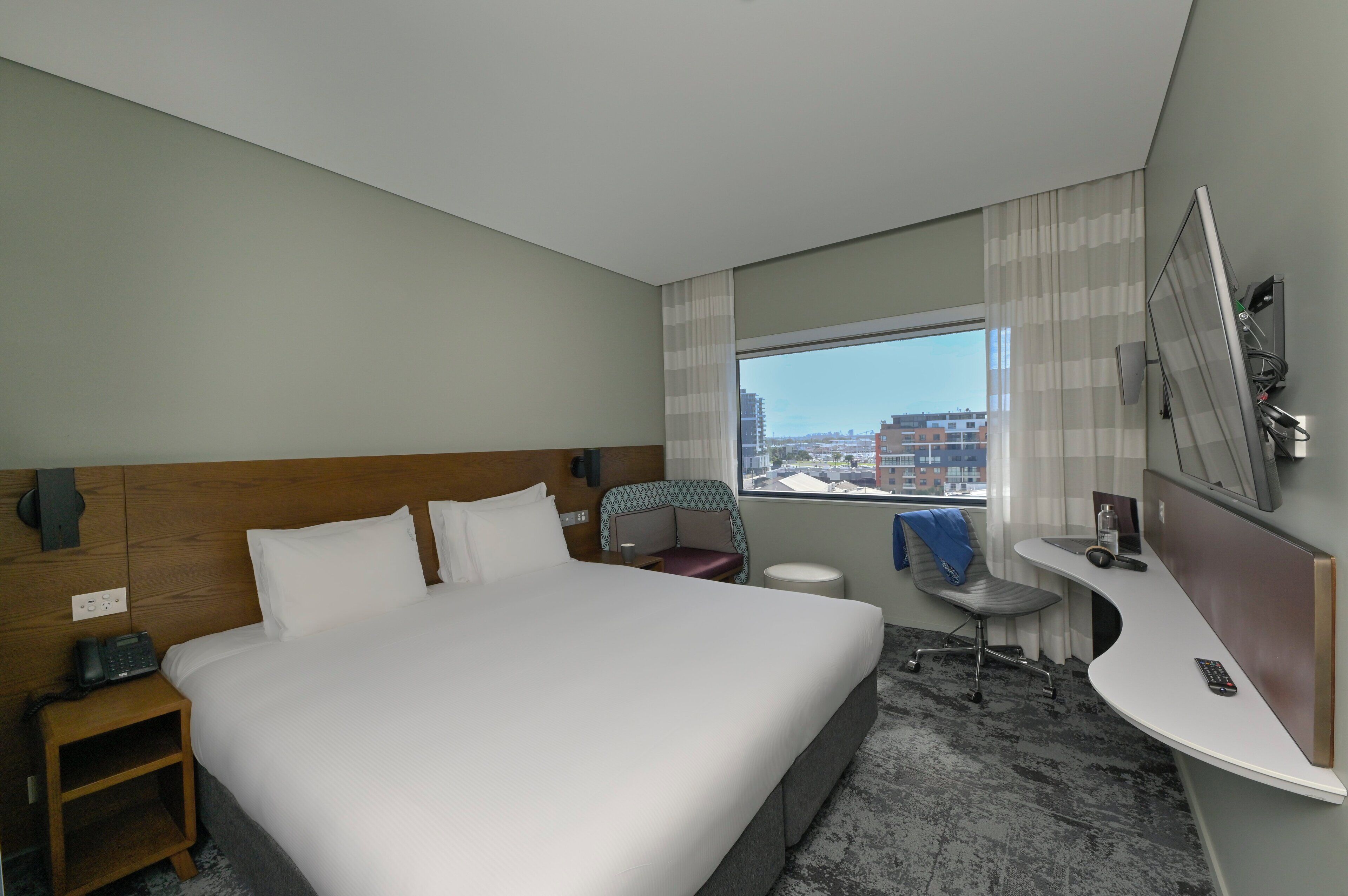 A hotel room with a large bed, a desk, a chair, a small table, and a window with a city view.
