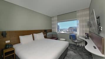 A hotel room with a large bed, a desk, a chair, a small table, and a window with a city view.
