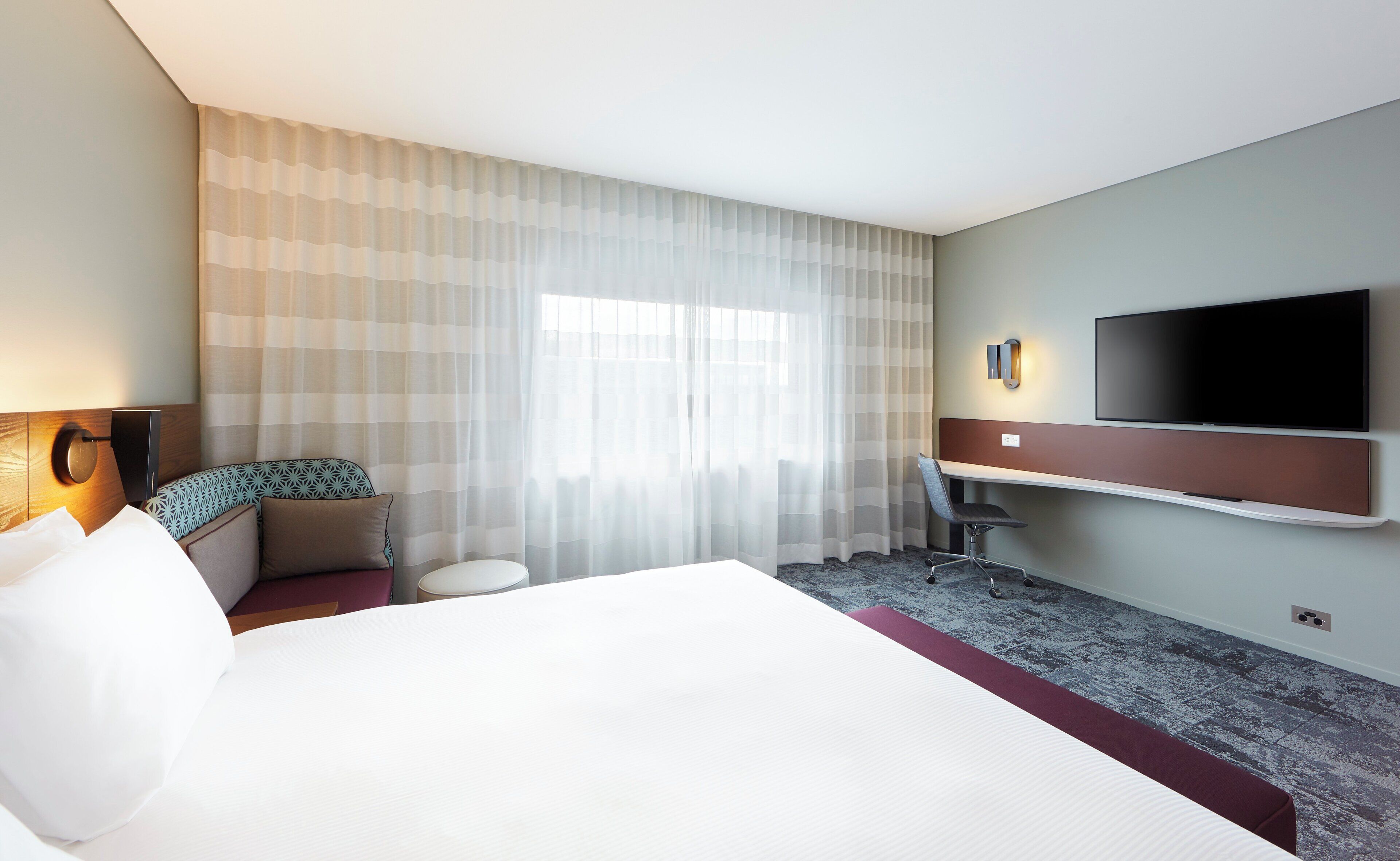 A hotel room with a bed, a desk, a chair, a TV, and a window with curtains.