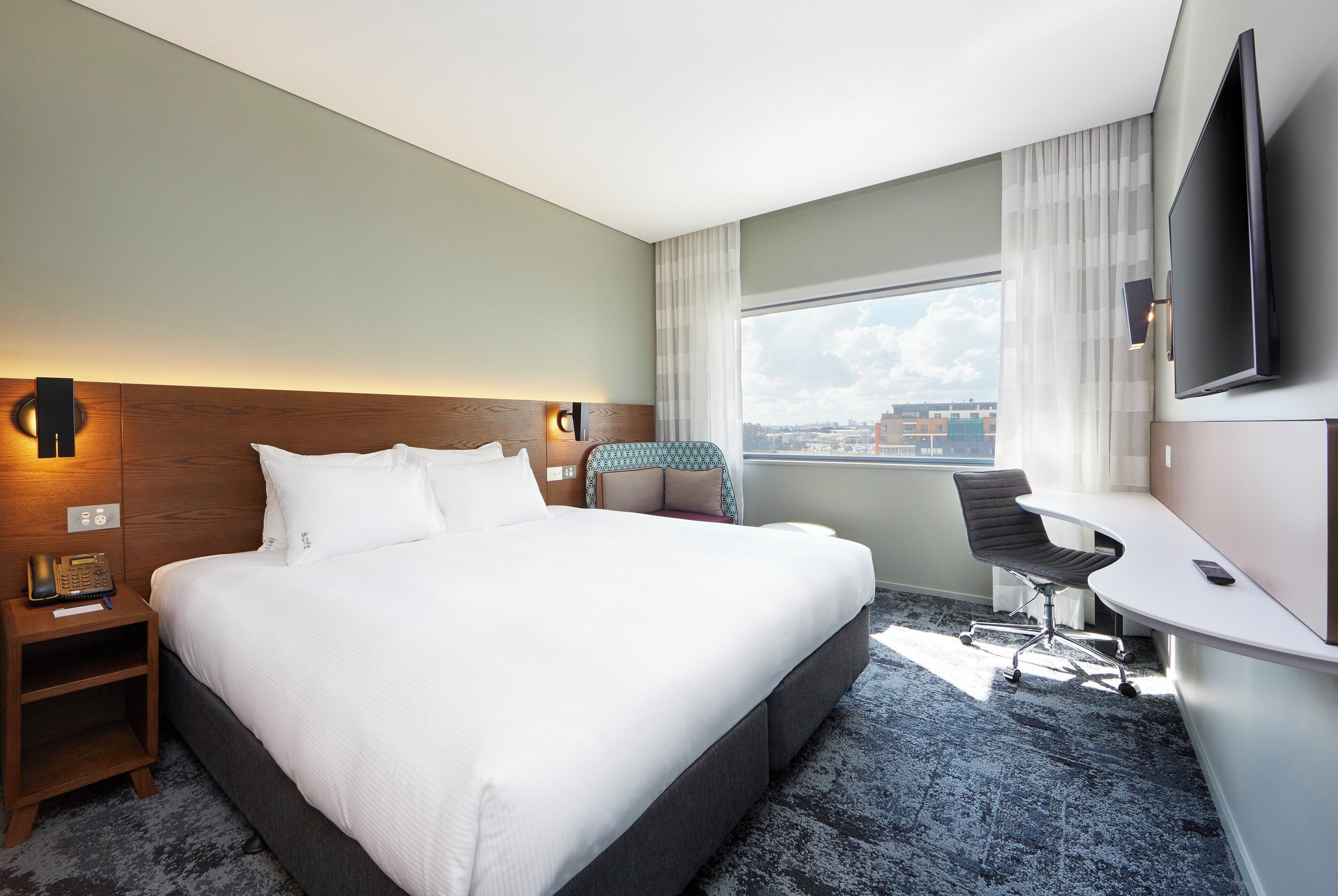A hotel room with a large bed, a desk, a chair, a TV, and a city view.