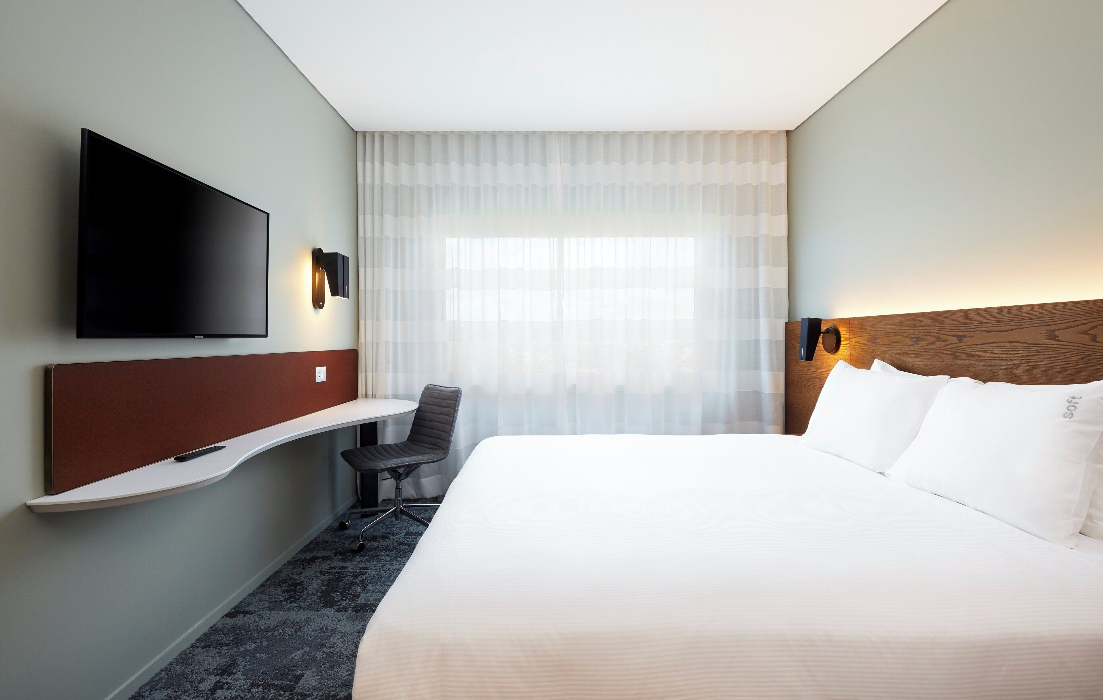A hotel room with a bed, a desk, a chair, a flat-screen TV, and a window with sheer curtains.