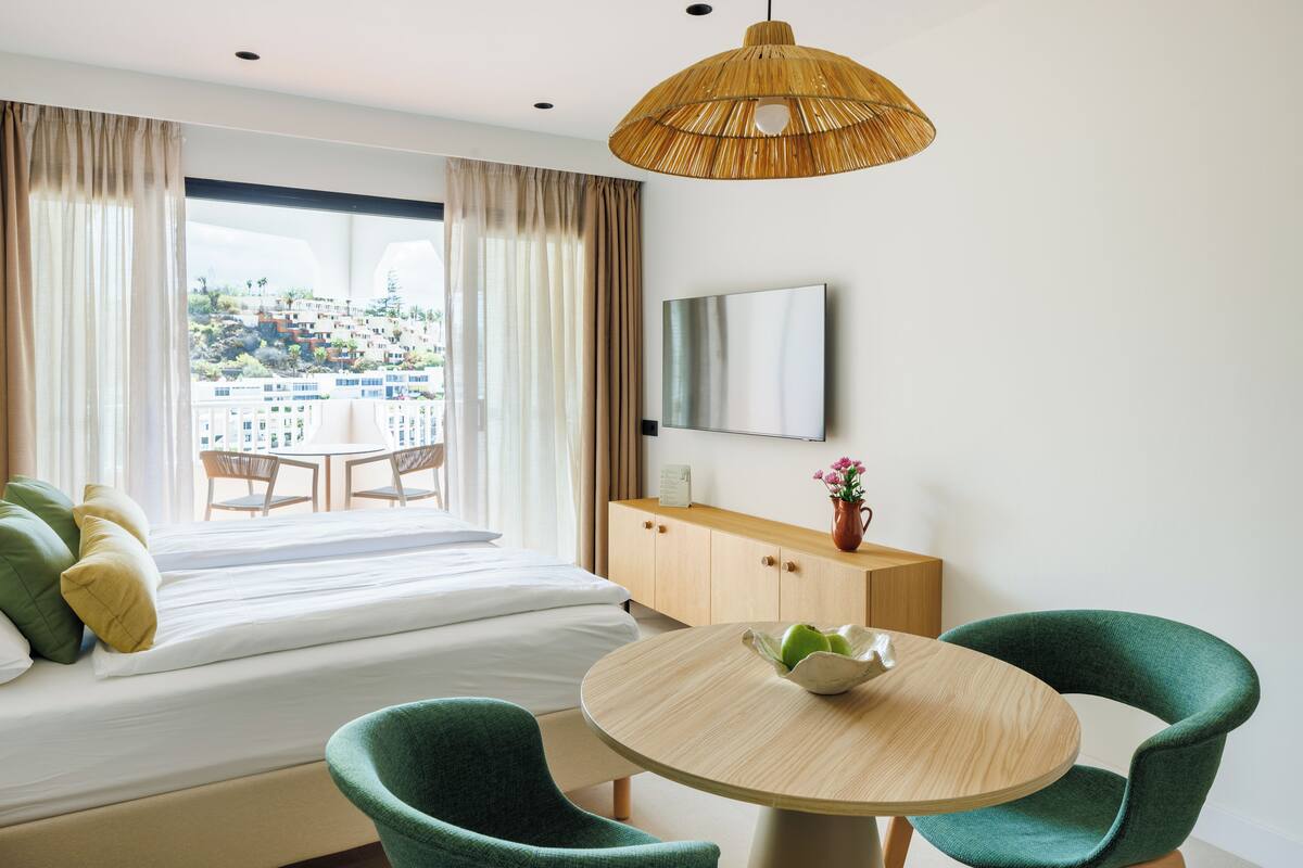 Ein Hotelzimmer mit Bett, Esstisch, zwei Stühlen, Fernseher und Blick auf eine Stadtlandschaft.