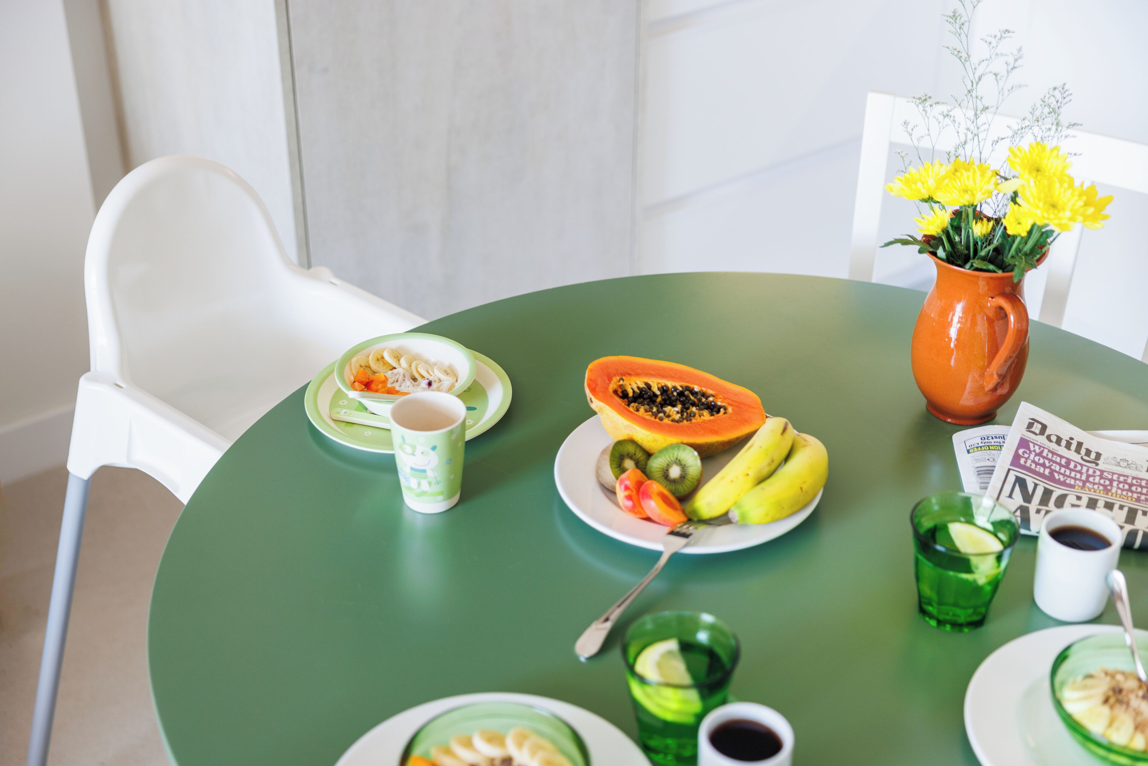 Una mesa con mantel verde, una silla blanca, un plato con fruta, un florero con flores amarillas, un periódico y un vaso de bebida verde.