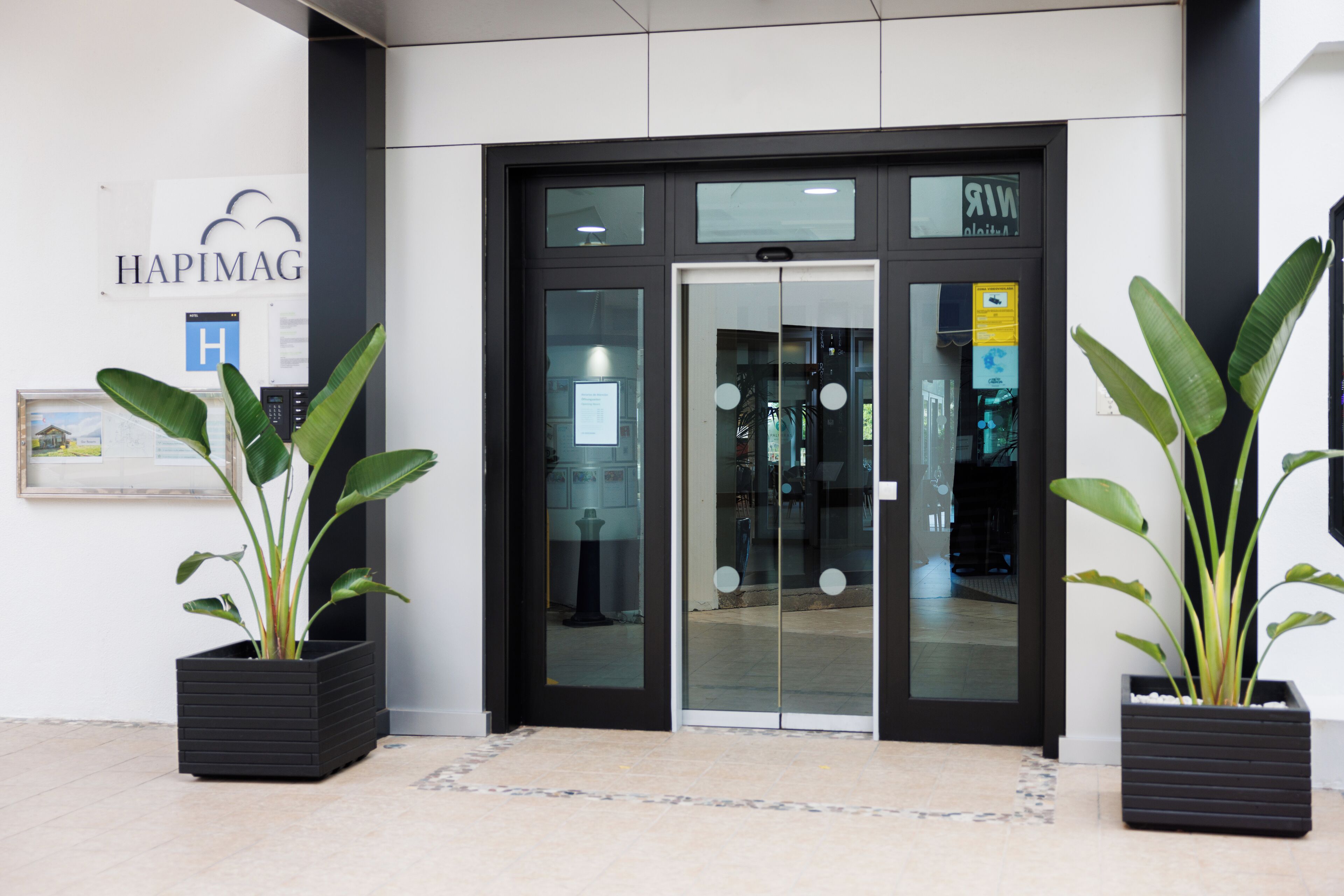 A modern building entrance with glass doors, a sign reading "HAPIMAG," and two potted plants.