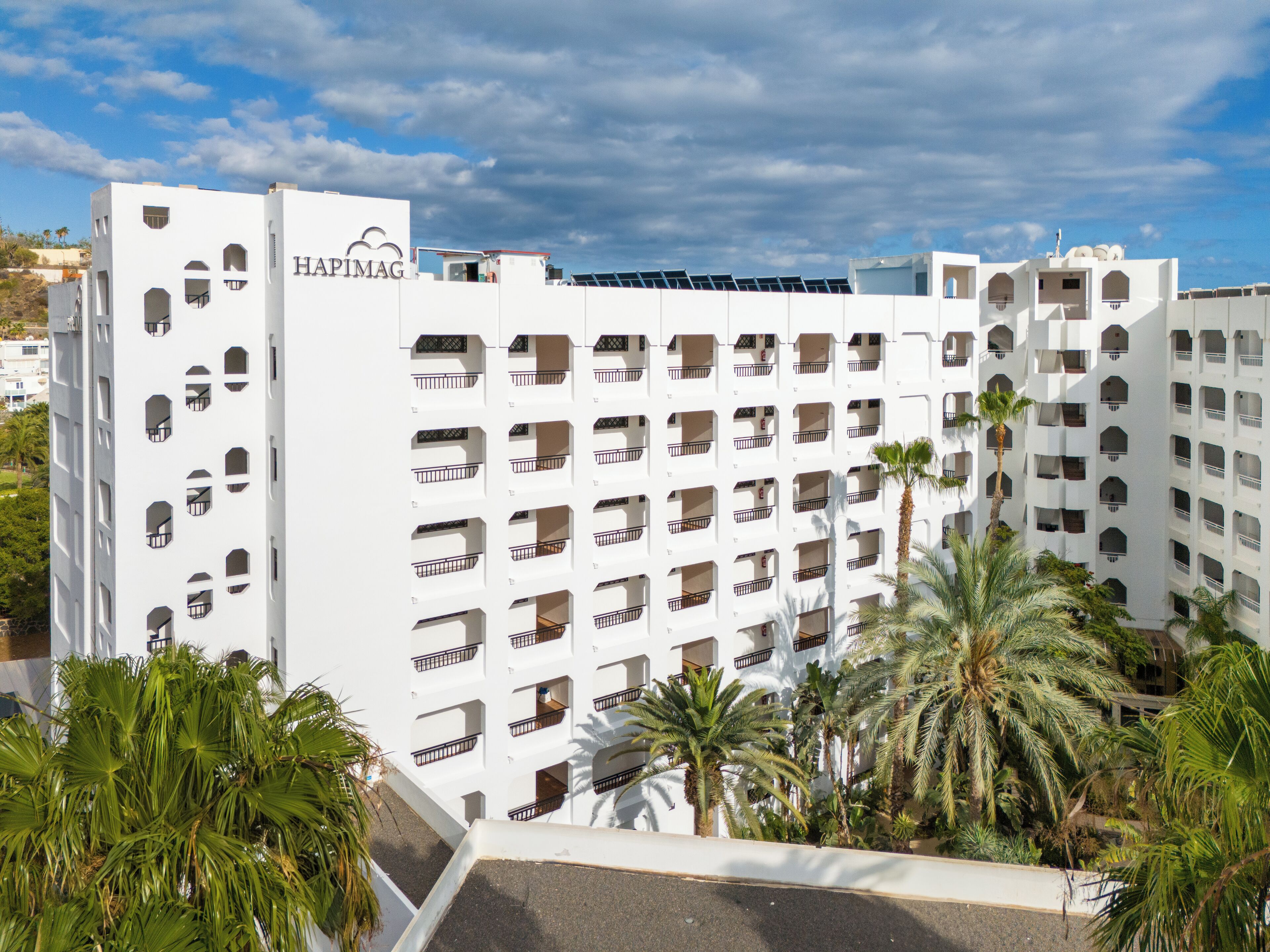 Un hotel de varios pisos con fachada blanca, numerosos balcones y palmeras en primer plano.