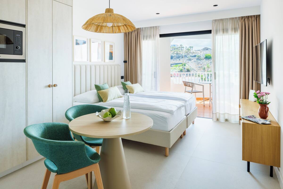 Ein modernes Hotelzimmer mit Bett, Esstisch mit Stühlen, Fernseher und Balkon mit Aussicht.
