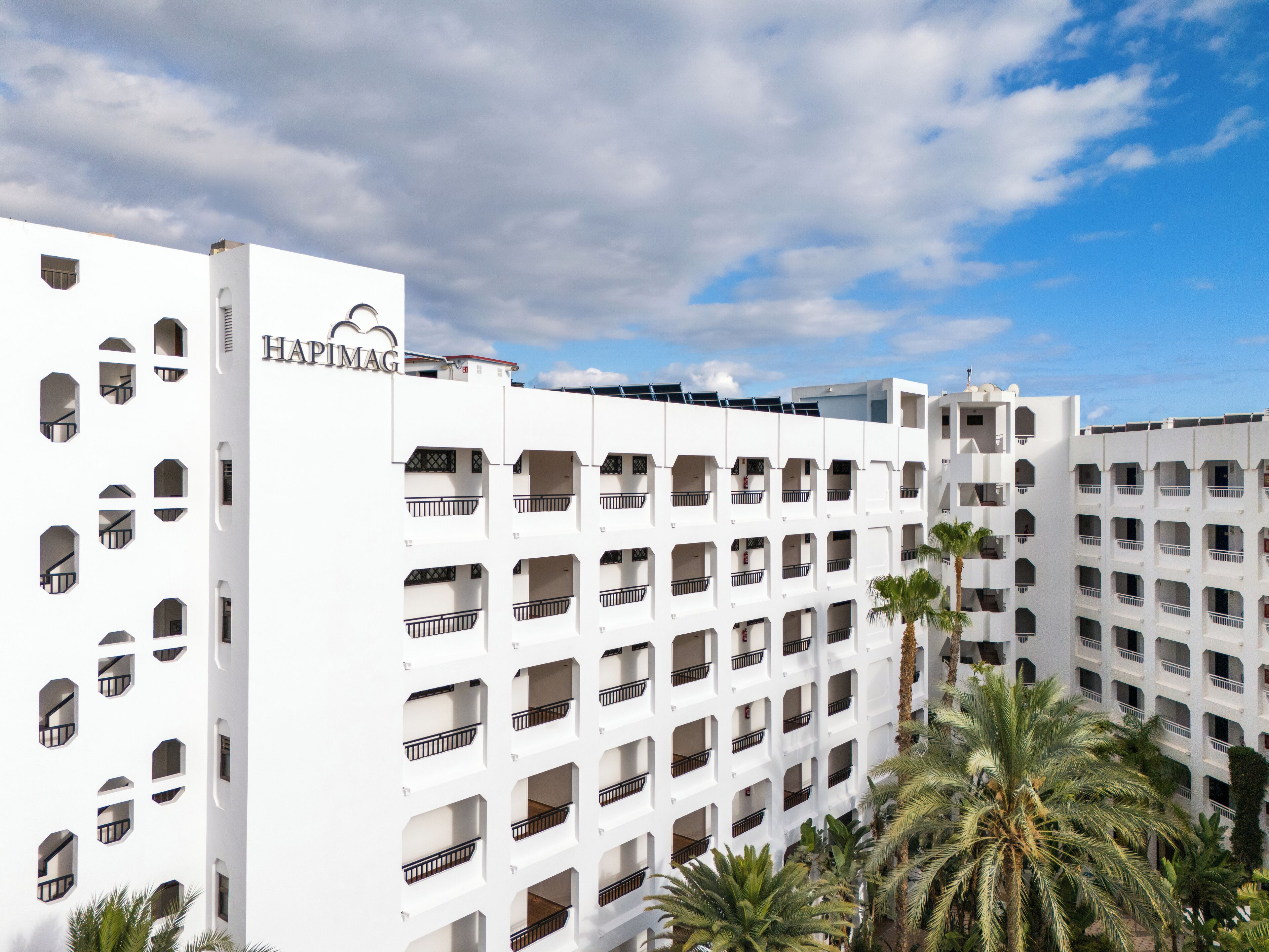 A multi-story hotel with a white facade, balconies, and palm trees in the foreground.