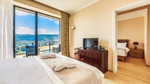 A hotel room with a large bed, a television, a desk with a phone, and a view of a city and mountains. - Lisi Hills Hotel (Tbilisi)
