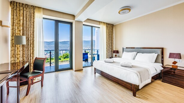 A hotel room with a bed, a desk, a chair, and a balcony with a view of the city. - Lisi Hills Hotel (Tbilisi)