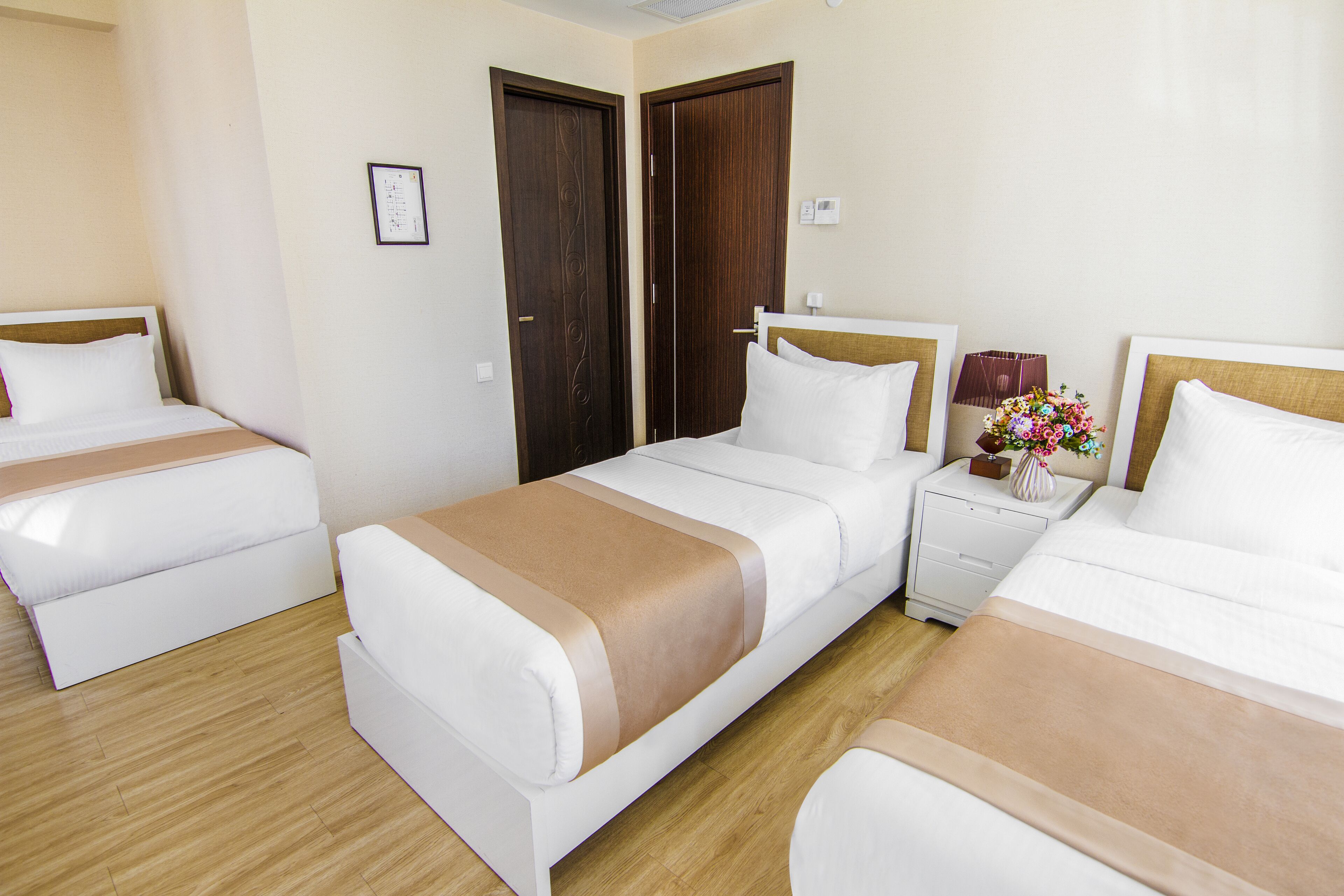 Habitación de hotel con dos camas individuales, una mesita de noche con lámpara y un jarrón de flores, y suelo de madera.