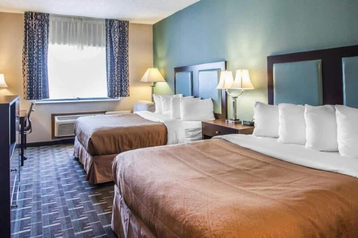 A hotel room with two beds, a desk, a chair, a window with curtains, and two bedside lamps.