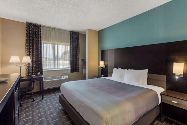 A hotel room with a large bed, a desk with a chair, a desk lamp, a nightstand, and a window with curtains.