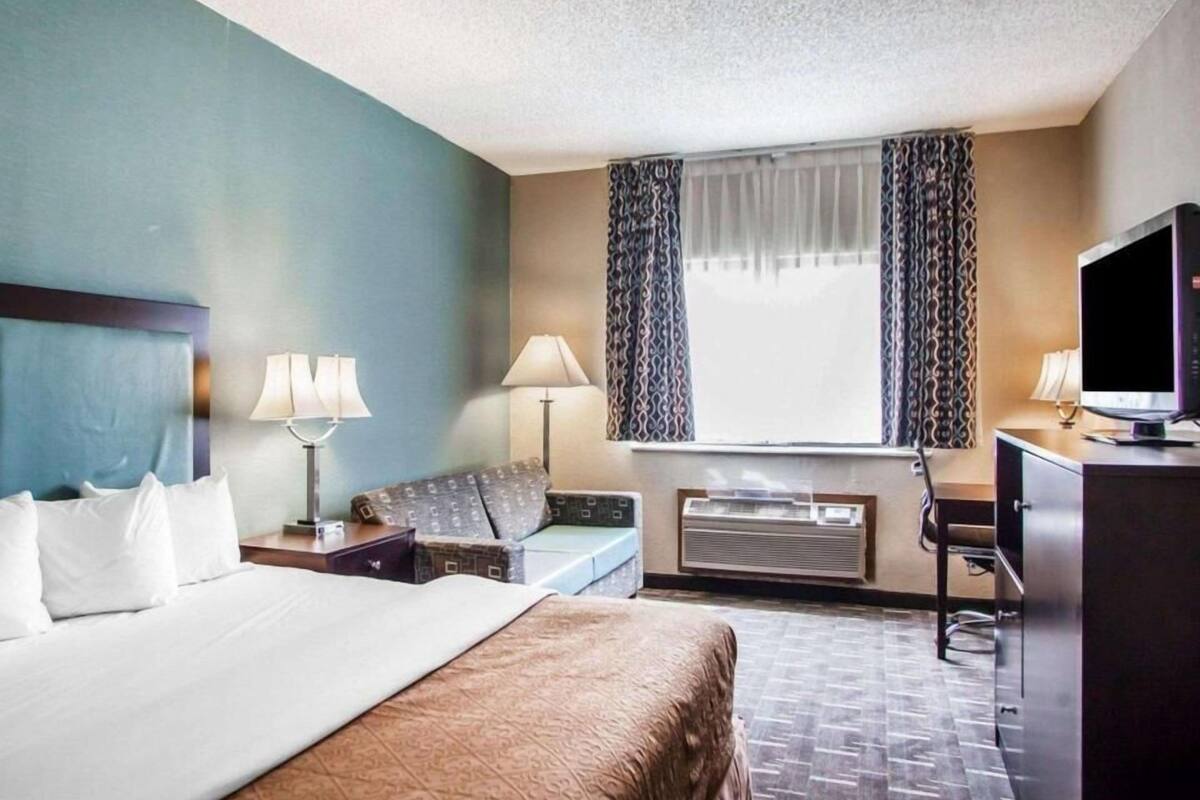 A hotel room with a bed, two bedside tables with lamps, a desk with a television, a window with curtains, and an air conditioning unit.