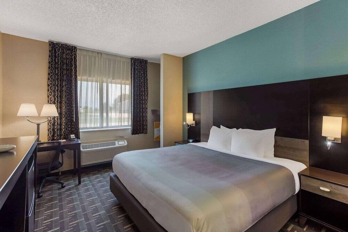 A hotel room with a large bed, a desk with a chair, a desk lamp, a nightstand, and a window with curtains.