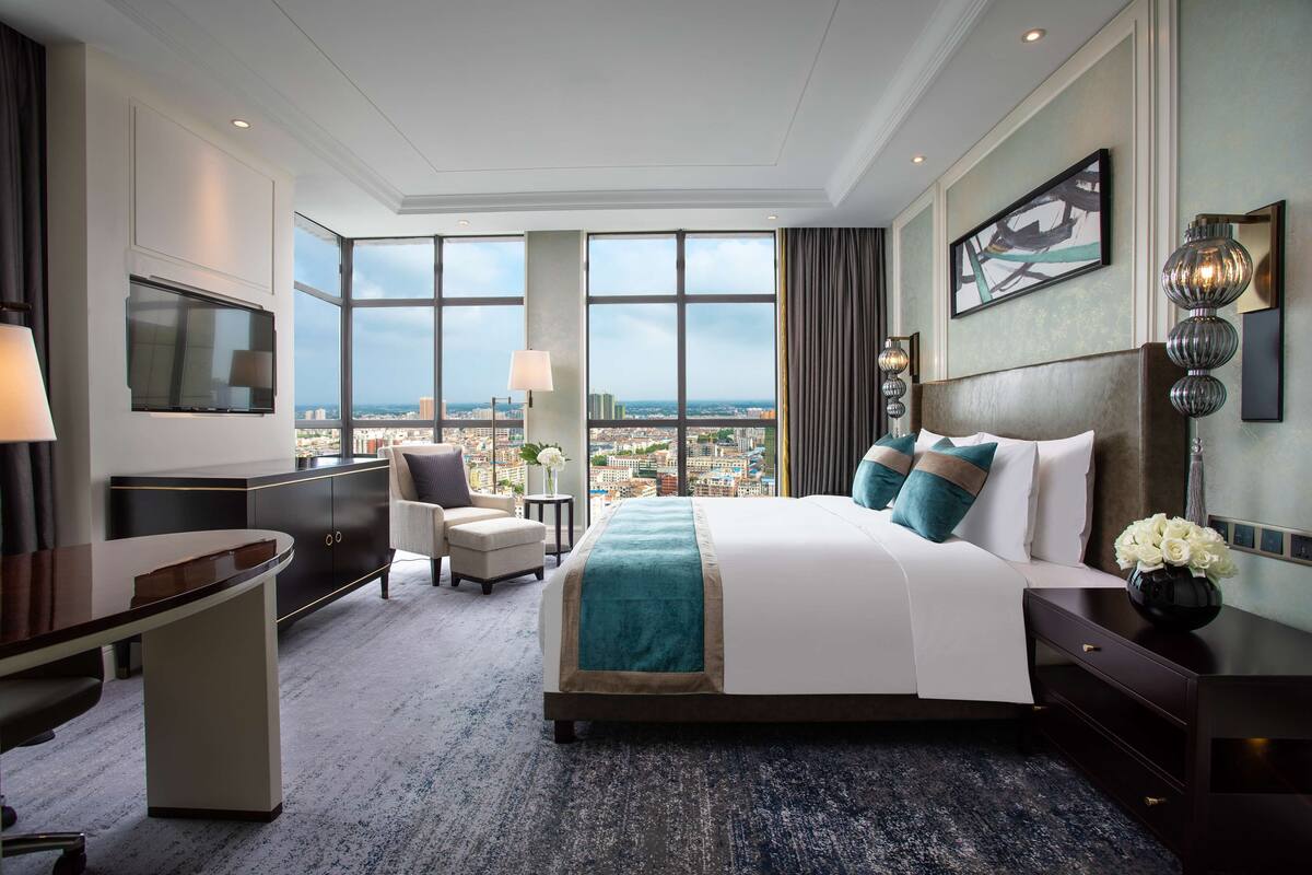 Une chambre d’hôtel moderne dotée d’un grand lit, d’un bureau, d’une chaise et offrant une vue sur la ville grâce à de grandes fenêtres.