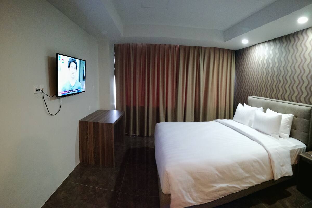 A hotel room with a bed, a TV mounted on the wall, a wooden nightstand, and a curtain.