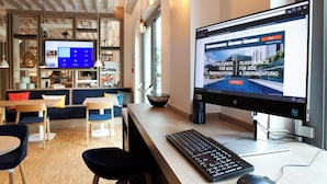 Un moderno vestíbulo de hotel con una pantalla de ordenador grande mostrando un sitio web, una zona de estar con mesas y sillas, y un amplio ventanal que proporciona luz natural.