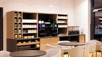A modern cafe with round tables and chairs, a coffee machine, and shelves stocked with mugs and condiments.