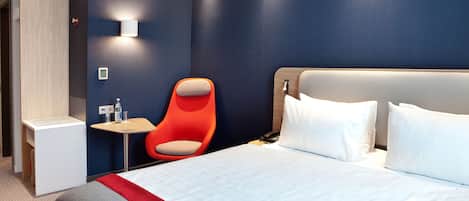Una habitación de hotel moderna con una cama grande, una silla roja, una mesita pequeña y una lámpara de pared.