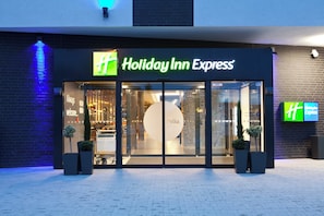 The exterior of a Holiday Inn Express hotel with a clear glass entrance, a 'Welcome' sign, and a 'Hello' sign inside.