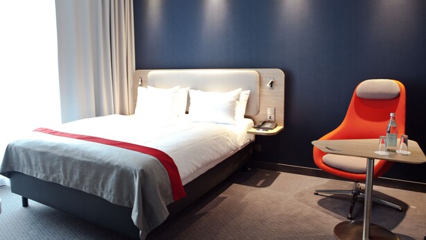 Una habitación de hotel con cama, mesita de noche, una silla roja y una pequeña mesa.