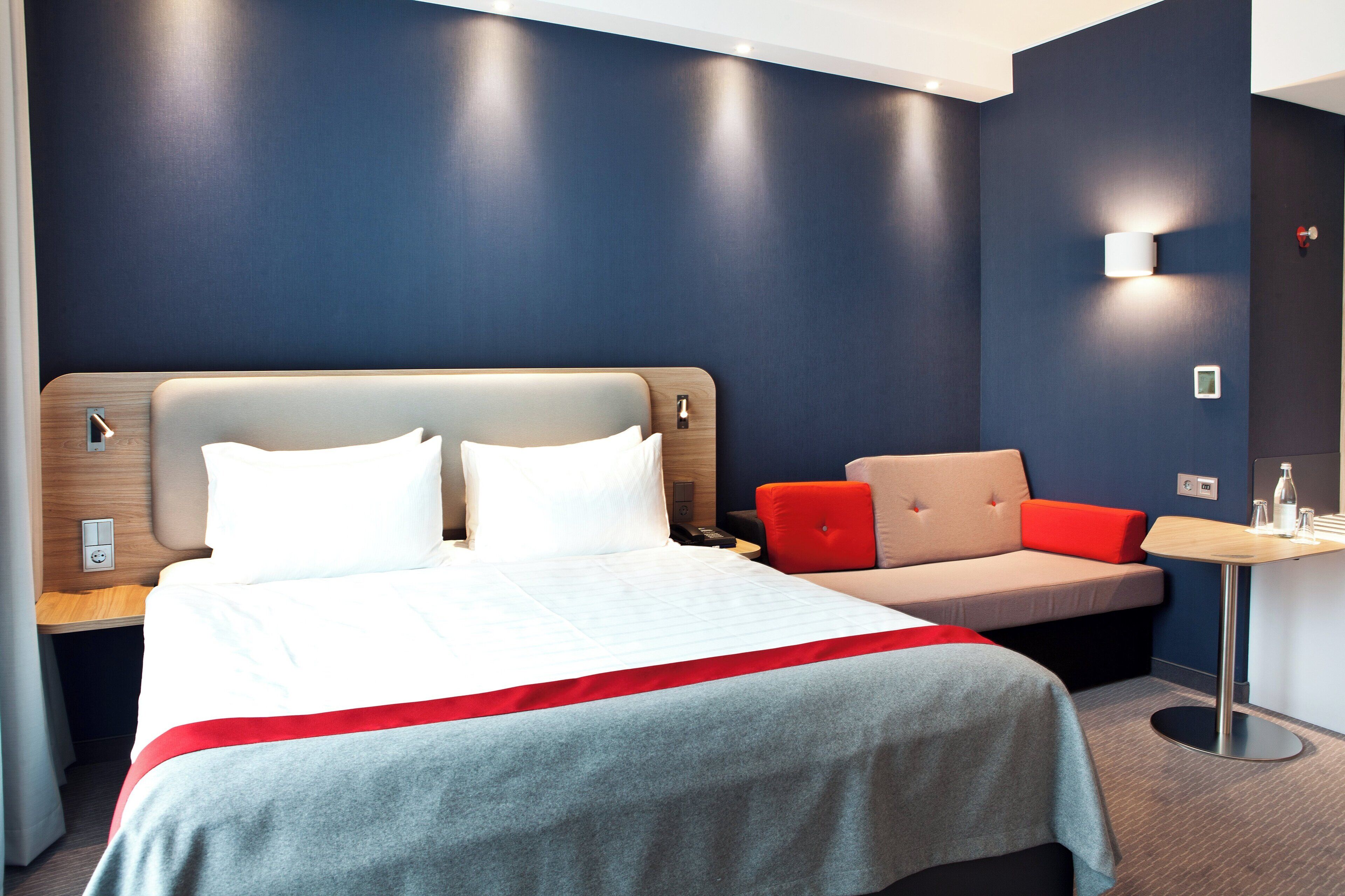A hotel room with a bed, a sofa, a small table, and a wall-mounted light.