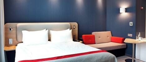A hotel room with a bed, a sofa, a small table, and a wall-mounted light.