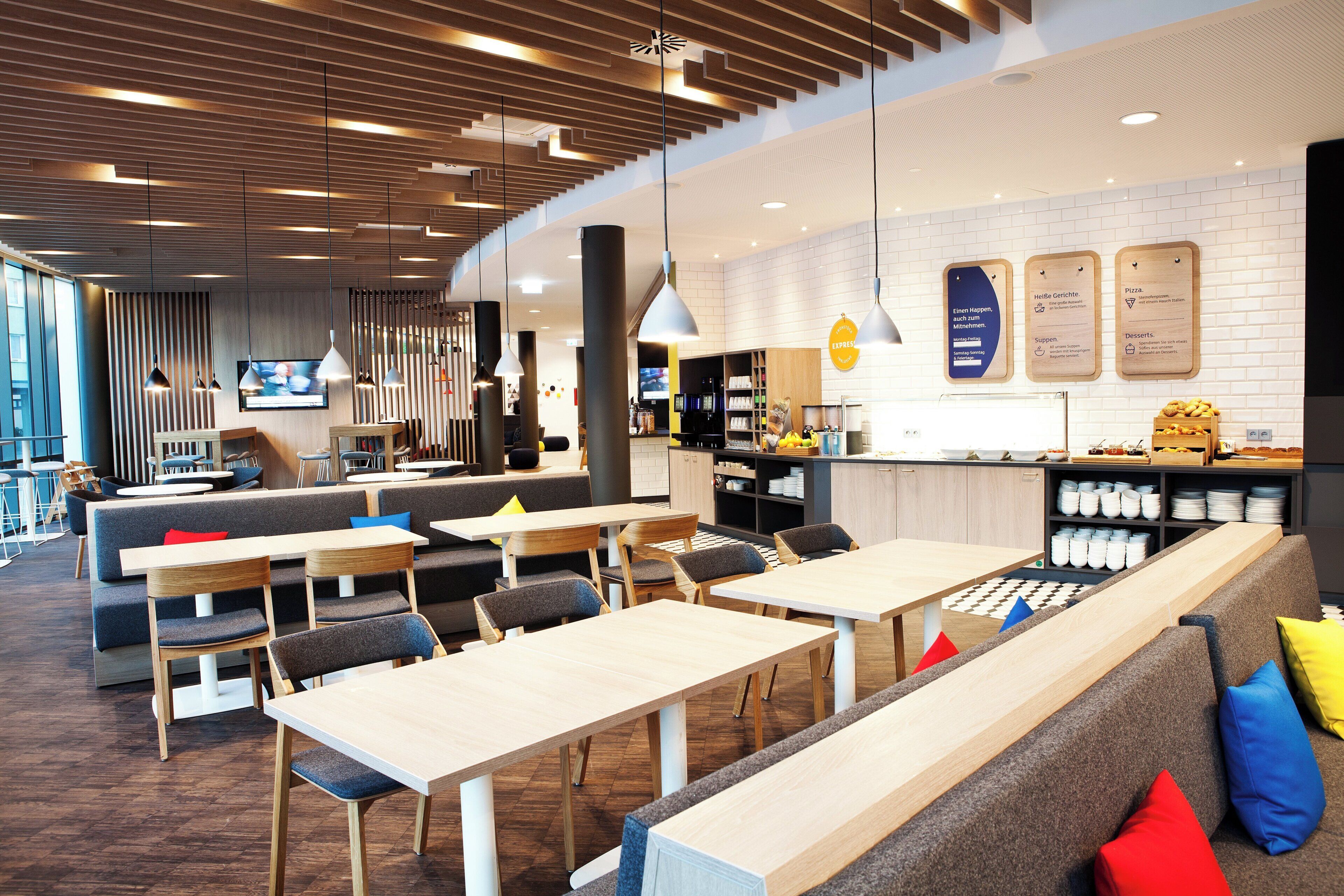 A modern restaurant interior with wooden tables and chairs, a counter with a menu, and a seating area with cushions.