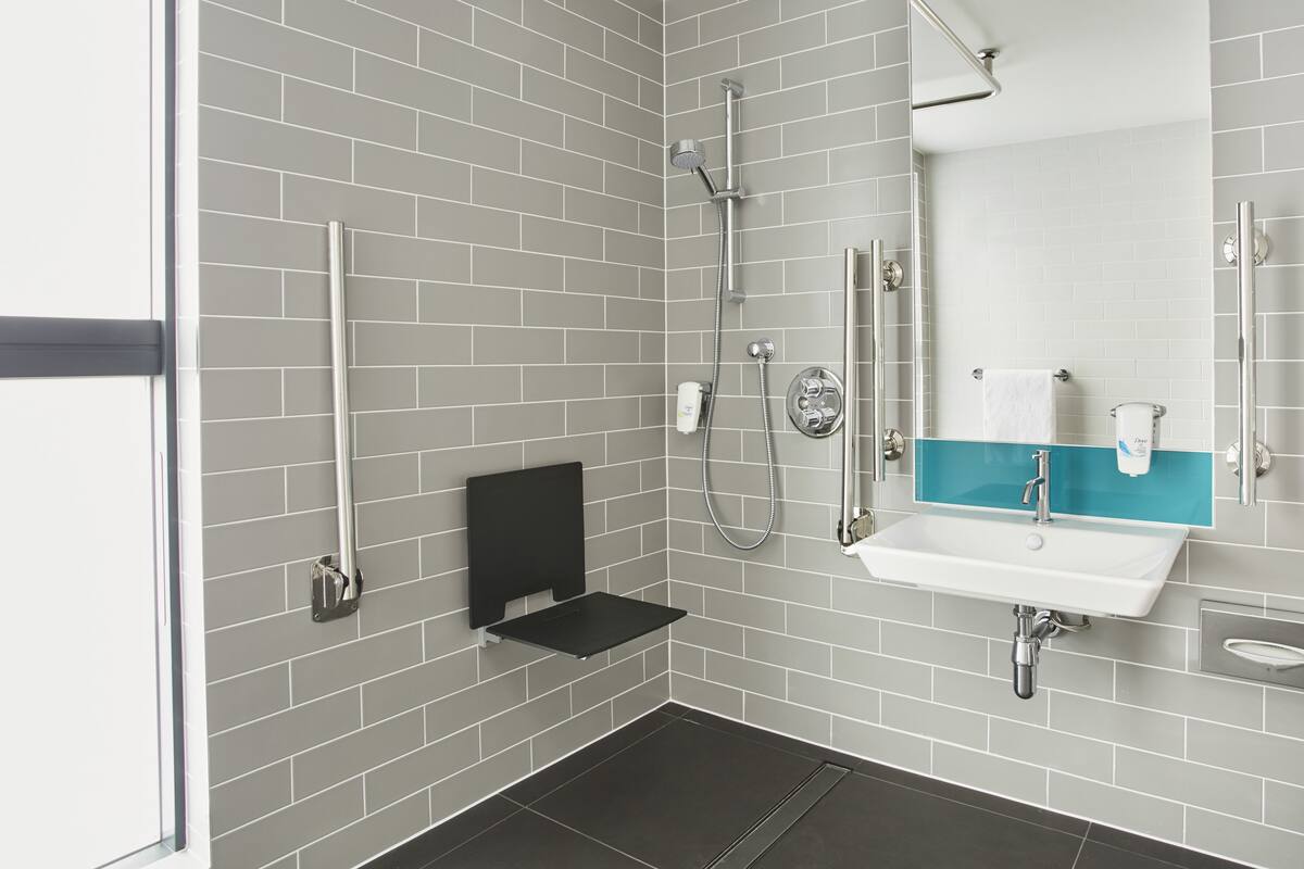 A modern bathroom with grey tiled walls, a black shelf, a white sink with a blue accent, and a glass shower enclosure.