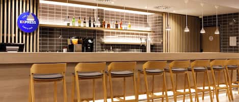 A modern bar area with a long counter, multiple bar stools, and a variety of bottles on the shelf.