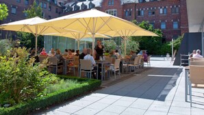 Un café al aire libre con mesas y sombrillas amarillas, rodeado de edificios de ladrillo y follaje.