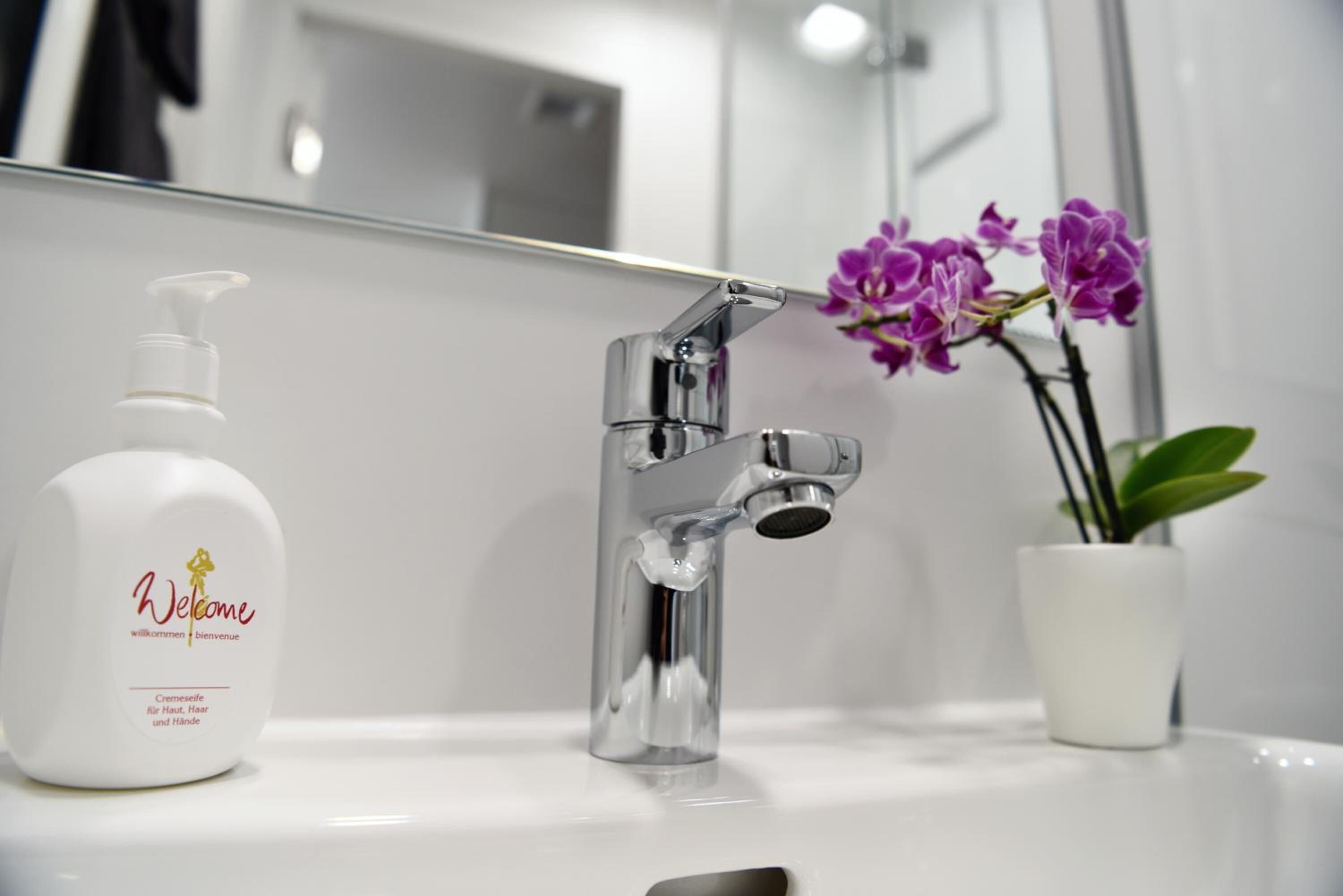 Ein Badezimmer mit einem chromfarbenen Wasserhahn, einer weißen Shampooflasche und einer Topf-Orchidee.