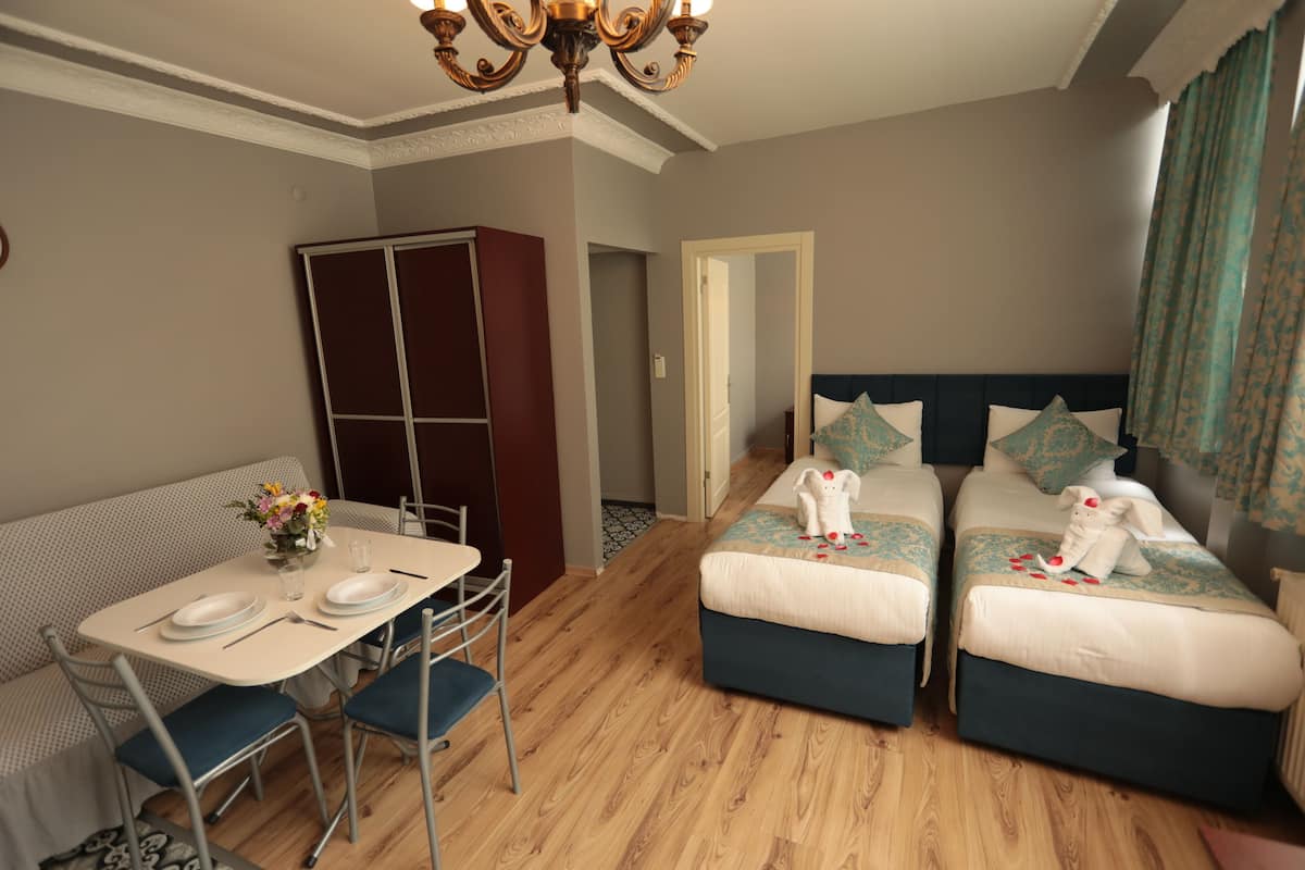 A room with a dining table set for two, two single beds with decorative pillows, a wardrobe, and a chandelier.