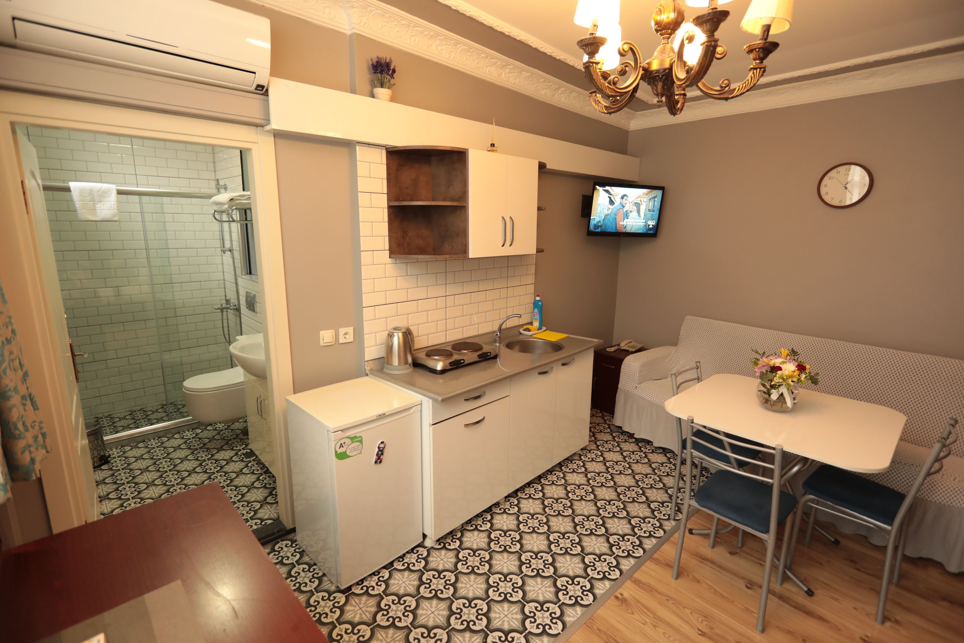 Cocina compacta con lavamanos, estufa y un refrigerador pequeño. Hay una zona de comedor con mesa y sillas, y un televisor montado en la pared.