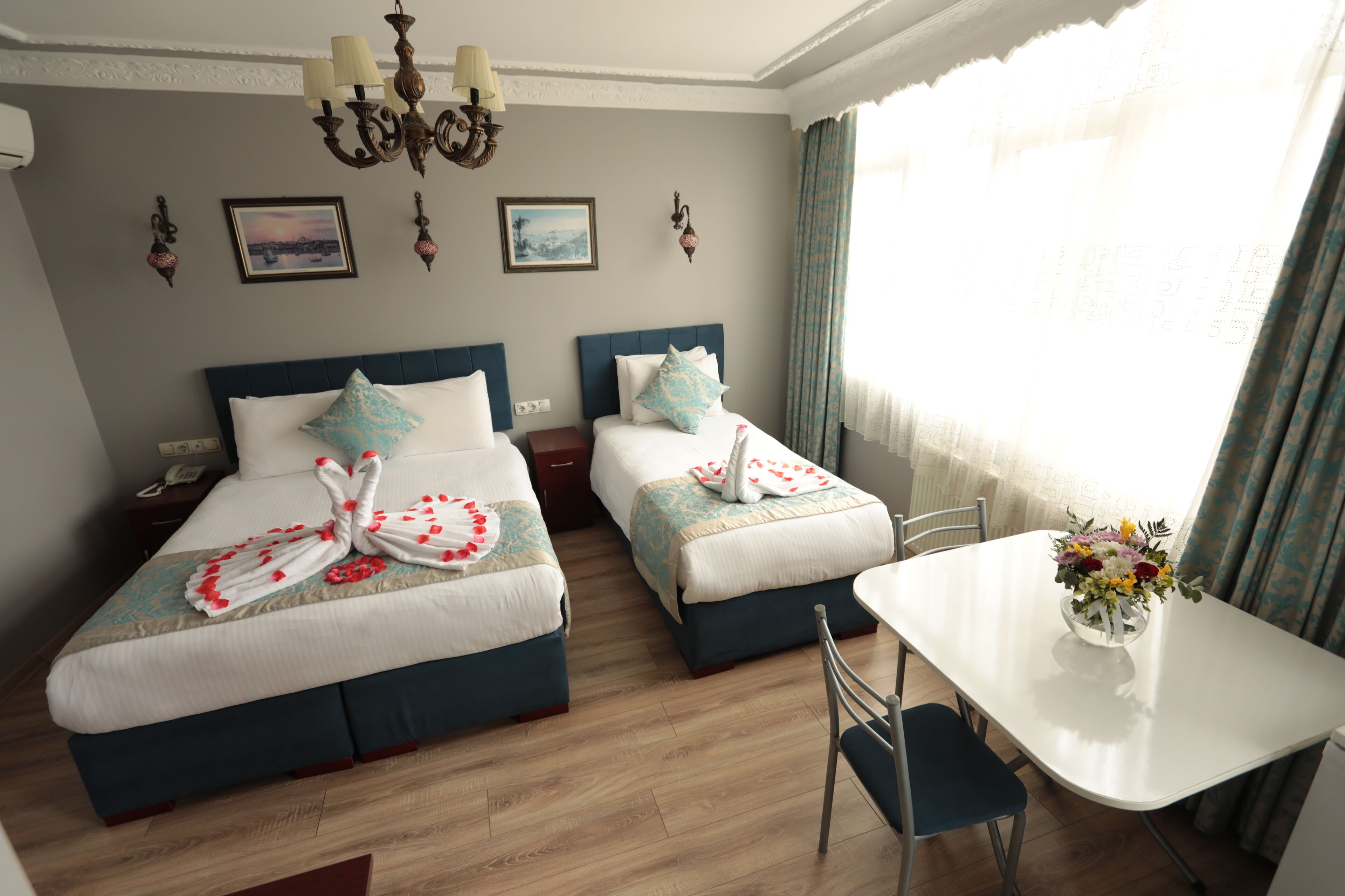 A hotel room with two beds, a dining table, a chandelier, and a window with curtains.