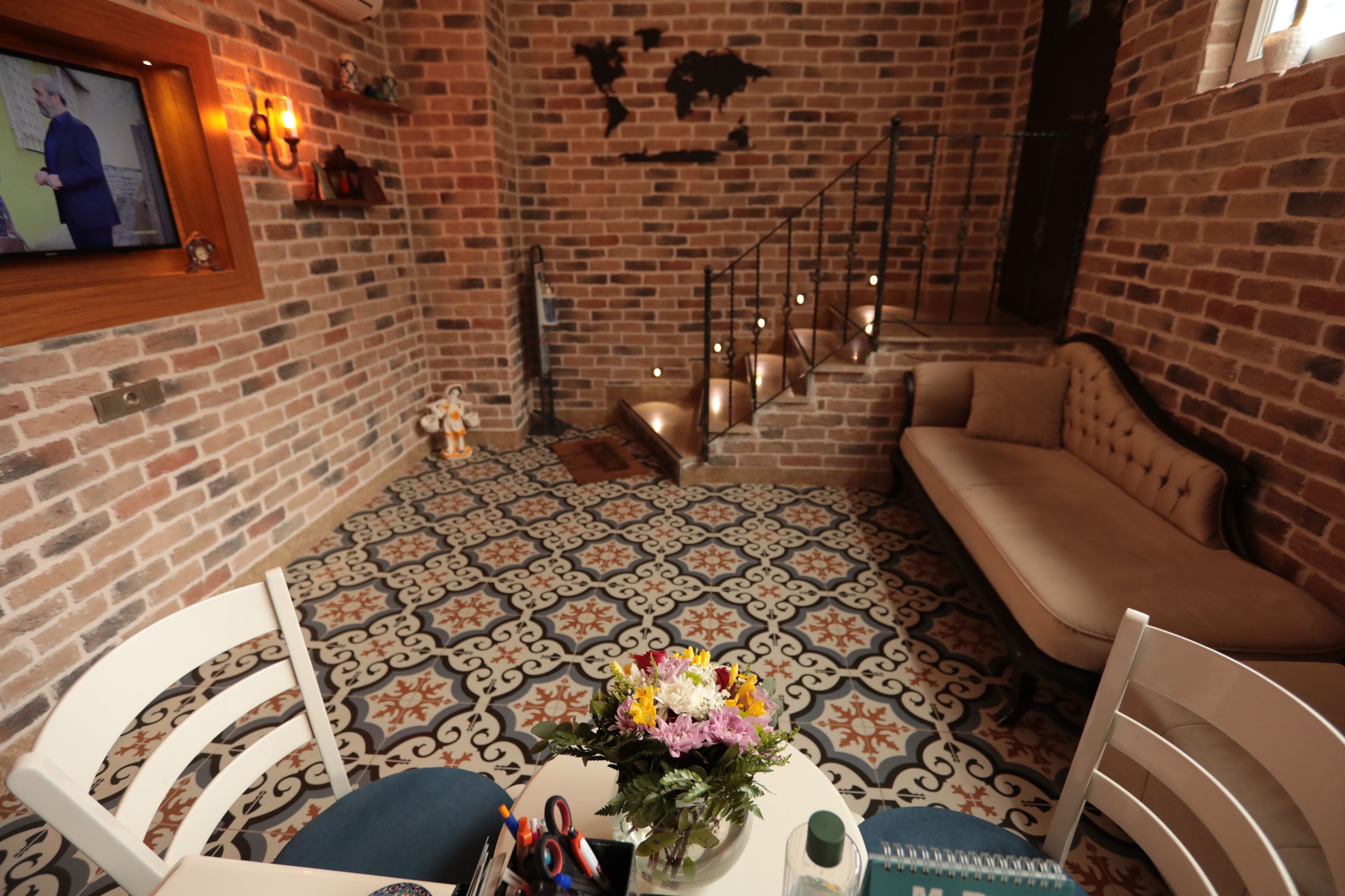 Ein gemütliches Zimmer mit Backsteinwand, einem gemusterten Boden, einem Sofa, einem Sessel und einem kleinen Tisch mit Blumenvase.
