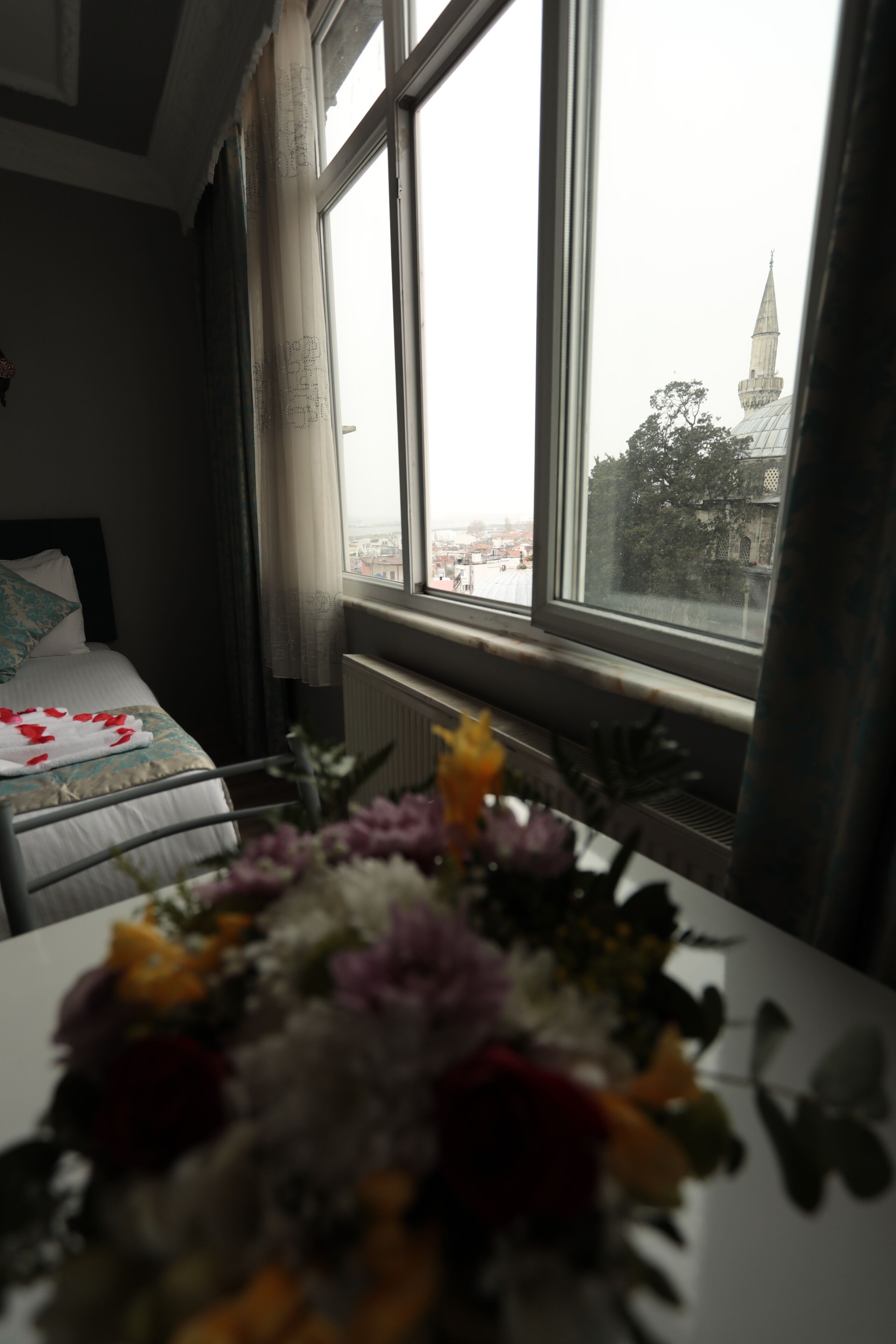 A hotel room with a bed, a window with a view of a building, and a table with a floral arrangement.