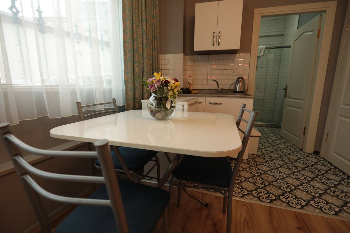 A dining area with a white table, blue chairs, and a vase with flowers. There is a kitchen in the background with white cabinets and a tiled backsplash.