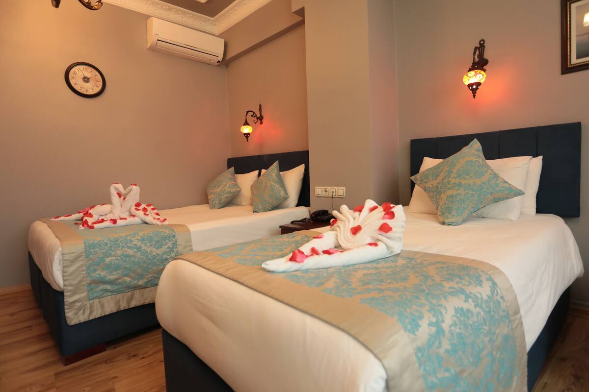 A hotel room with two beds, each with a blue and white patterned bedspread, a white and blue pillow, and a towel arranged on the bed. There is a clock on the wall and a wall-mounted light fixture.