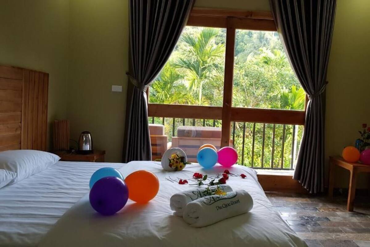 A hotel room with a bed, a window with curtains, a small table with a vase and balloons, and a wooden headboard.