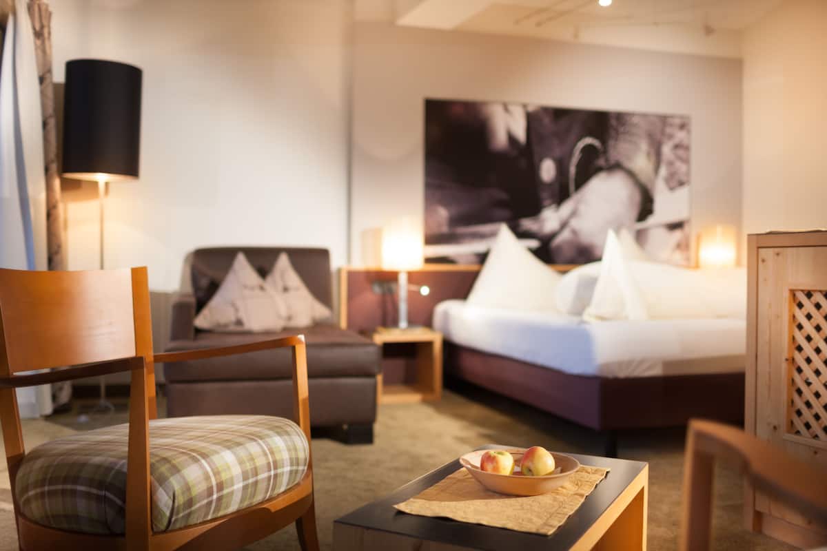 A hotel room with a bed, a sofa, a chair, a table with a bowl of fruit, and a painting on the wall.
