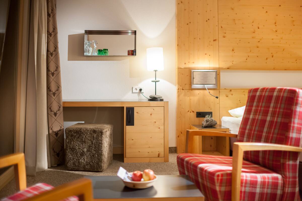 A hotel room with a wooden desk, a red plaid chair, a small bedside table, and a lamp.
