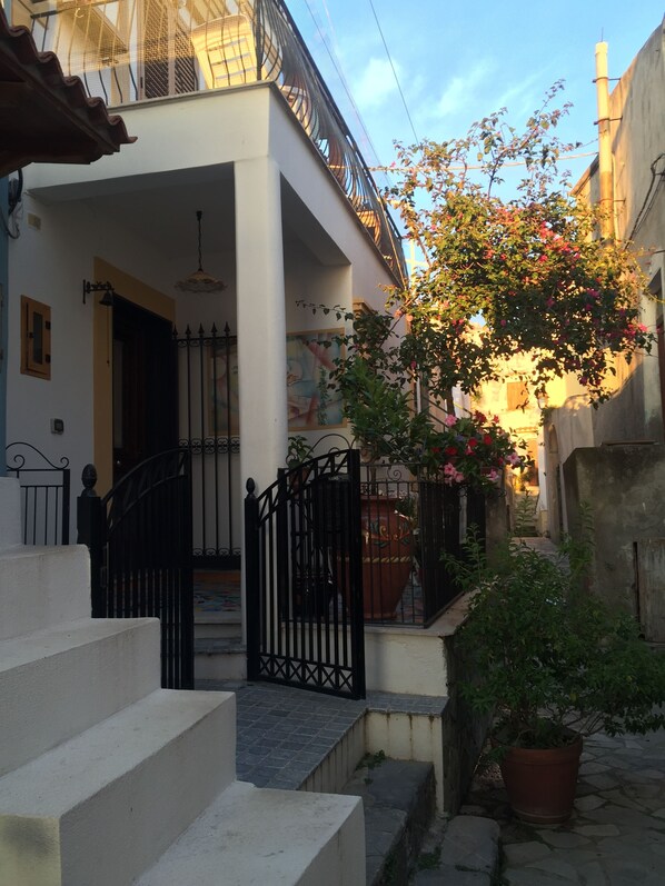 A narrow courtyard with a white building, a black gate, and a potted plant with pink flowers.