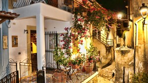 A narrow street with a building featuring a balcony with pink flowers, a staircase, and a stone wall.