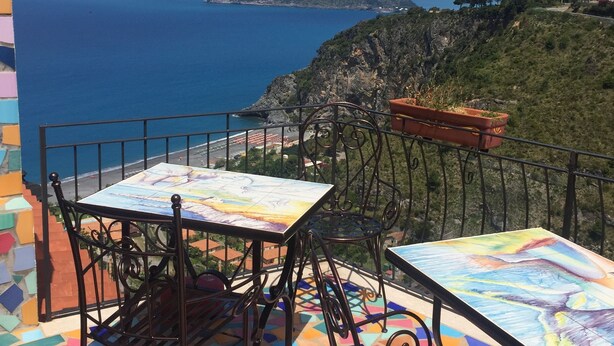 A terrace with two tables and chairs, a colorful tiled floor, and a view of the sea and a distant island.